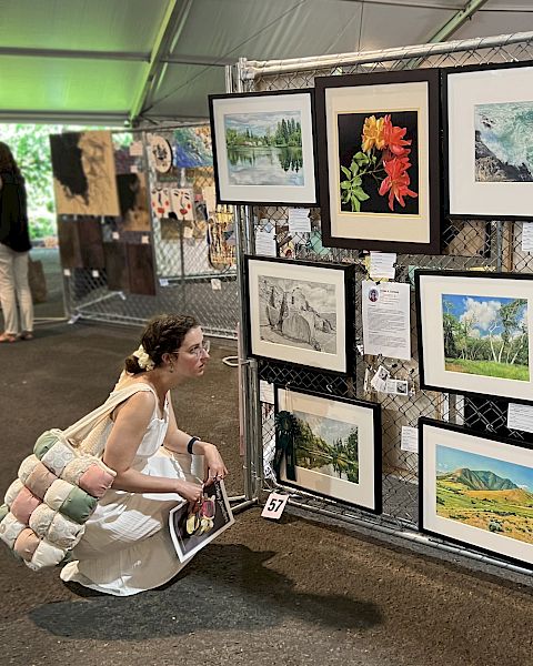 A woman kneels to view framed landscape and nature paintings at an art exhibition.
