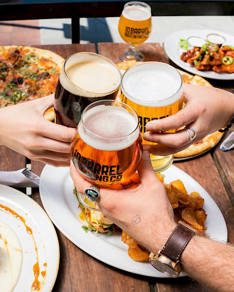 Friends clink beer glasses over a table of pizza, fries, and burgers, sharing a meal outdoors in a casual, messy, delicious feast.