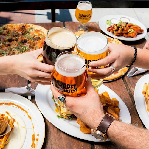Friends clink beer glasses over a table of pizza, fries, and burgers, sharing a meal outdoors in a casual, messy, delicious feast.