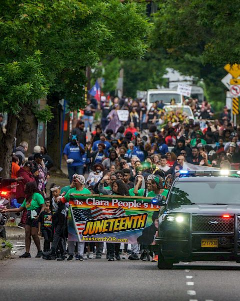 Protesters march down a city street with a banner &ldquo;ONE TENY&rdquo;? and a pursuing police SUV, crowds on both sides, trees lining the road. Ending with a period.