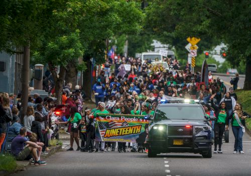 Protesters march down a city street with a banner &ldquo;ONE TENY&rdquo;? and a pursuing police SUV, crowds on both sides, trees lining the road. Ending with a period.