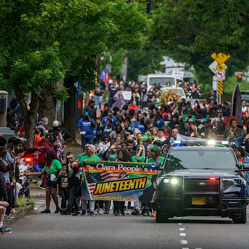 Protesters march down a city street with a banner &ldquo;ONE TENY&rdquo;? and a pursuing police SUV, crowds on both sides, trees lining the road. Ending with a period.