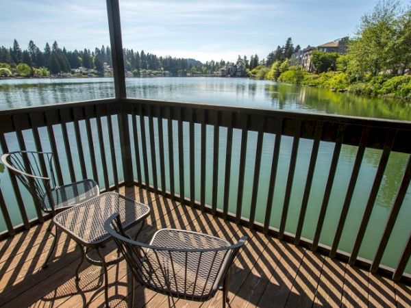 A wooden deck overlooks a calm lake with two metal chairs casting striped shadows, surrounded by green trees under a bright, clear sky.