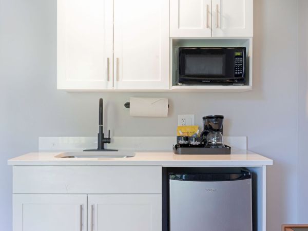 A compact kitchenette with white cabinets, a small sink, a microwave, a coffee maker, and a mini fridge under a countertop. End the sentence.