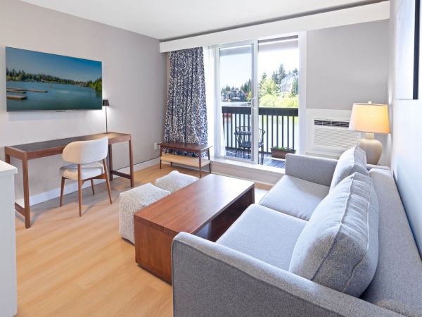 A bright, modern living room with a gray sofa, wooden coffee table, wall-mounted TV, a desk with chair, and a balcony view with curtains.