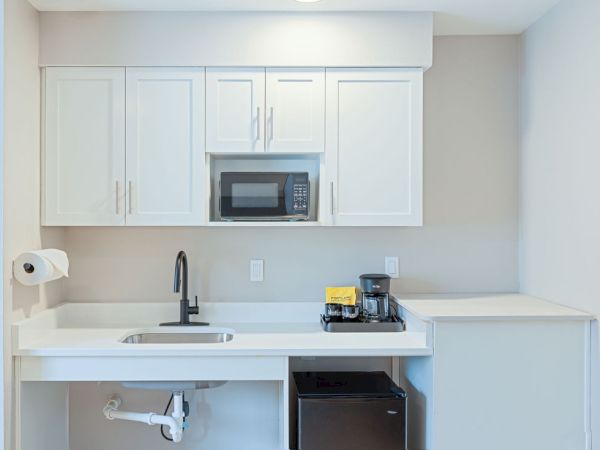 A compact, white kitchen with upper cabinets, a microwave, a small sink, a black faucet, a coffee maker, mini fridge, and a paper towel holder, all neatly arranged.