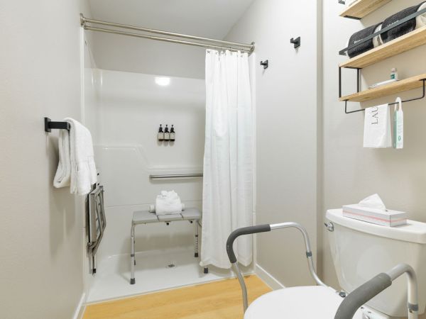 A compact accessible bathroom with a walk-in shower, grab bars, a raised toilet, a foldable shower bench, towel, and shelves.