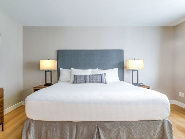 A neatly made hotel bed with a gray upholstered headboard, white linens, decorative pillows, two bedside lamps, and simple wooden furniture.