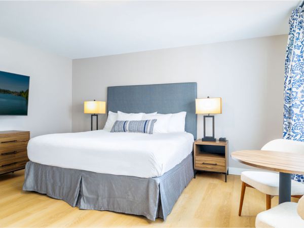 A bright hotel room with a king bed, gray headboard, two bedside lamps, a wooden dresser, a wall TV, and a small round table with chairs by a window with blue patterned curtains; modern and clean.