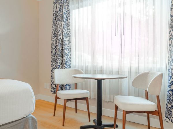 A hotel room corner with a round table and two white chairs by a large window, patterned curtains, and a bed partially visible to the left.