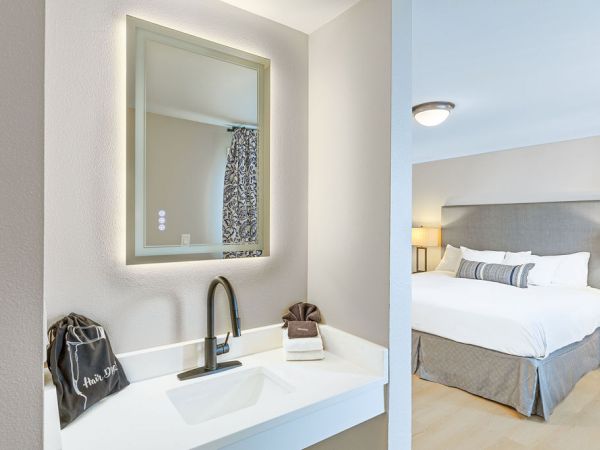 A modern hotel room with a small vanity area, wall mirror, and a bed visible through a doorway; clean, neutral decor with soft lighting.