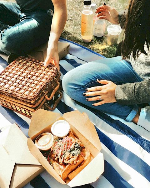 People having a picnic on a striped blanket with food boxes, drinks, and a woven basket.