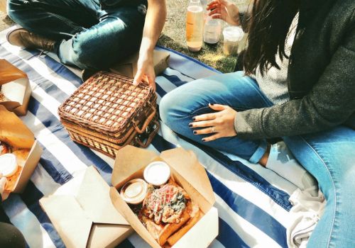 People having a picnic on a striped blanket with food boxes, drinks, and a woven basket.