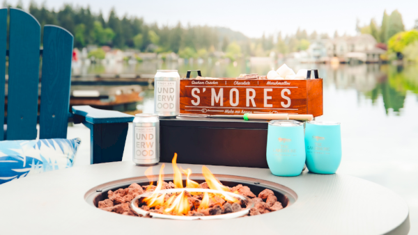 Outdoor fire bowl on a patio table by a river, with beer cans, cups, and a S&rsquo;mores box for roasting treats.