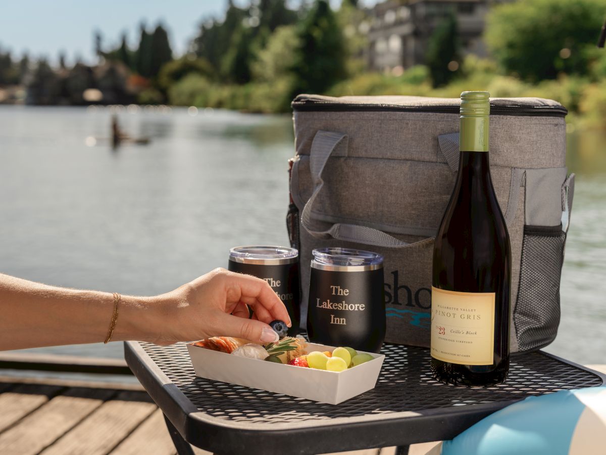 A riverside picnic setup: a cooler bag with wine bottle, two tumbler glasses, and snacks on a table by the lake.
